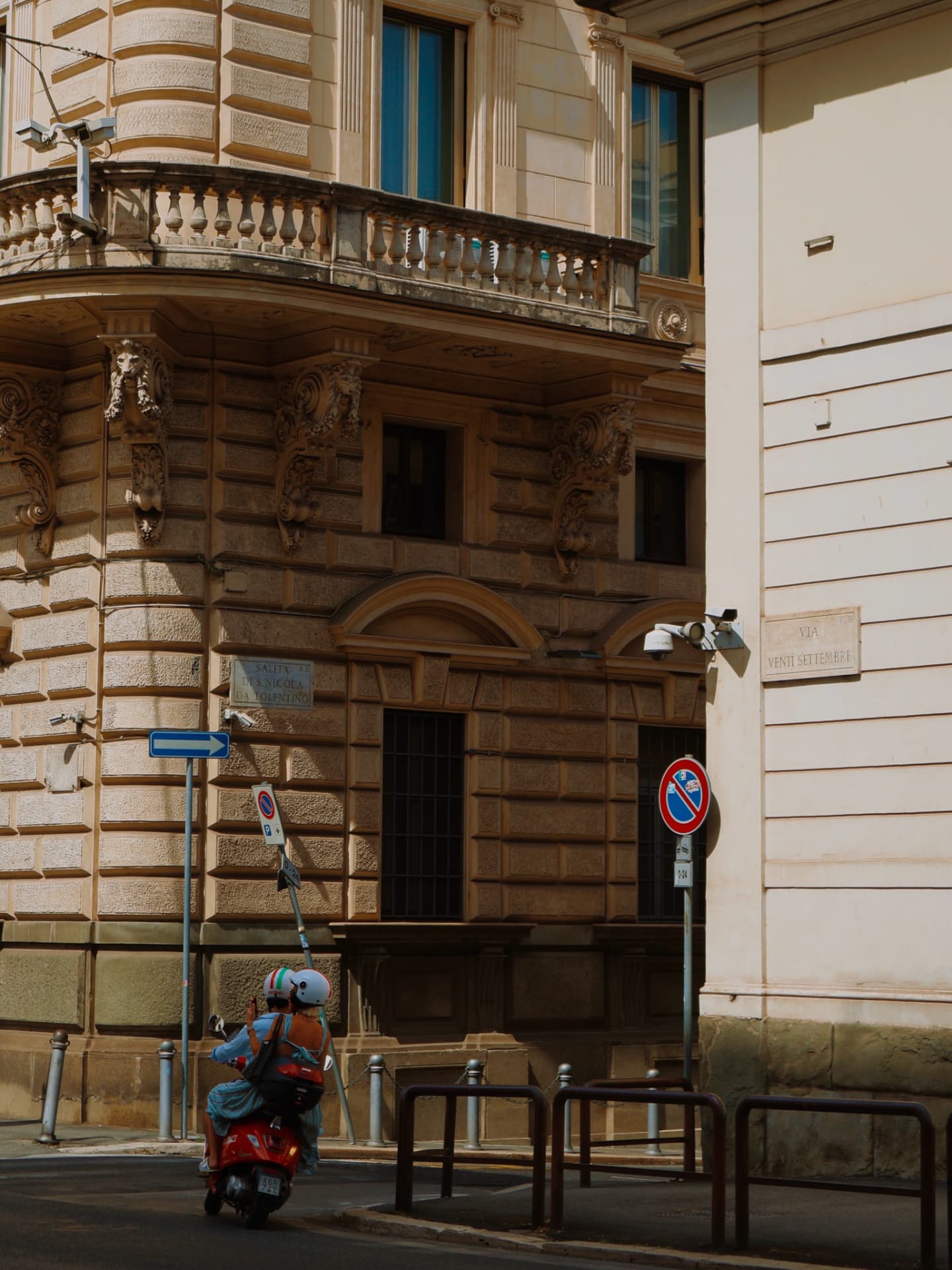 Two helmeted riders paused on a red scooter at a sunlit Roman street corner