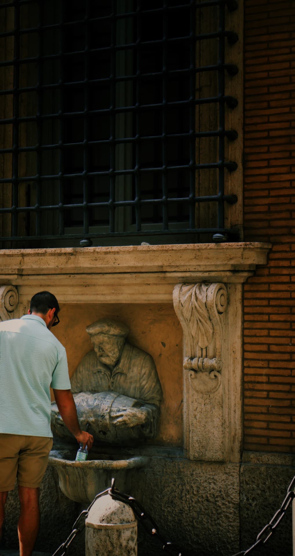 A man filling a water bottle at an old stone drinking fountain watched by a carved figure
