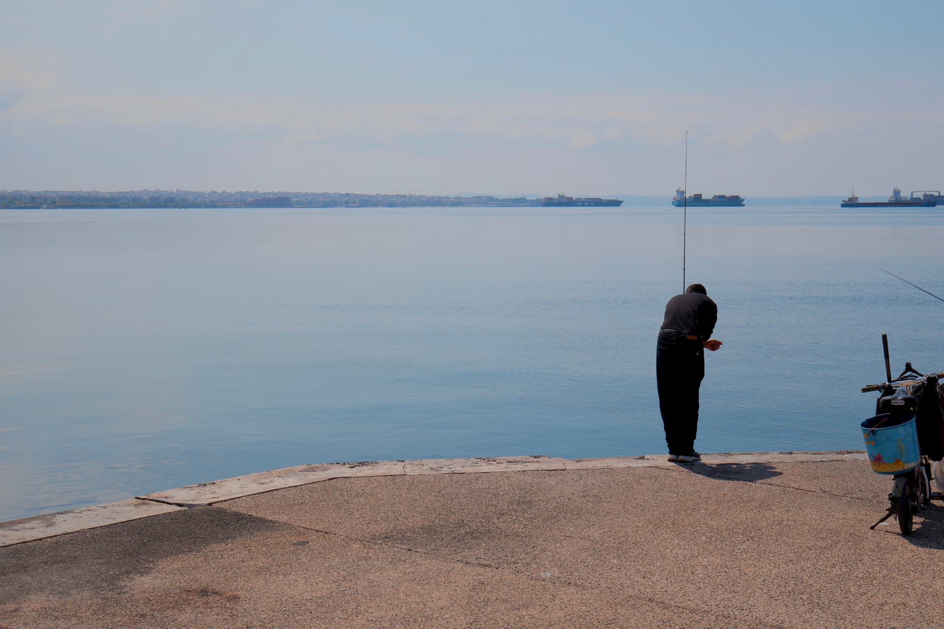 A fisherman standing at the edge of a stone promenade, cargo ships on the hazy horizon