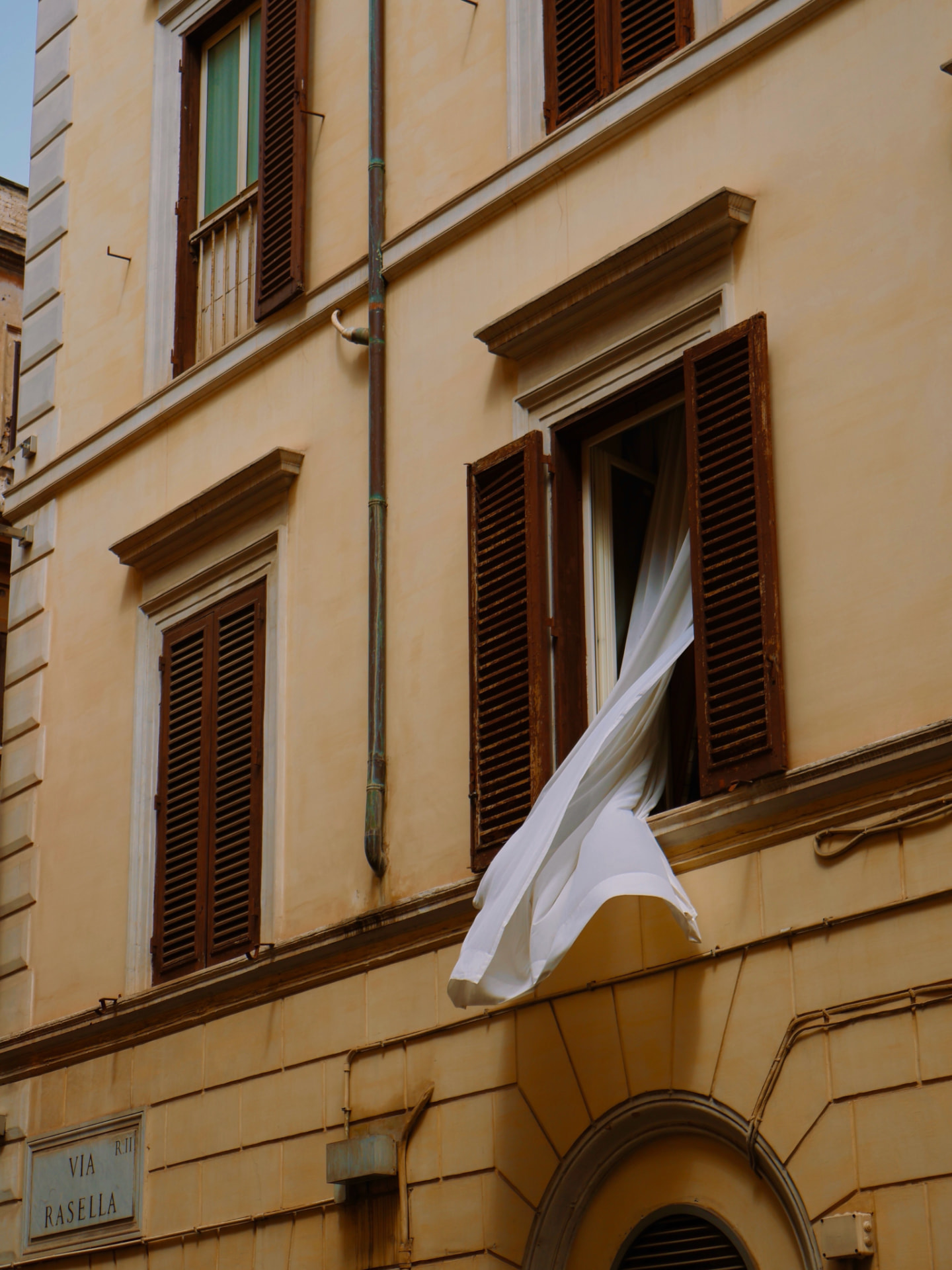 A sunlit Roman facade with wooden shutters and a white curtain caught in the breeze