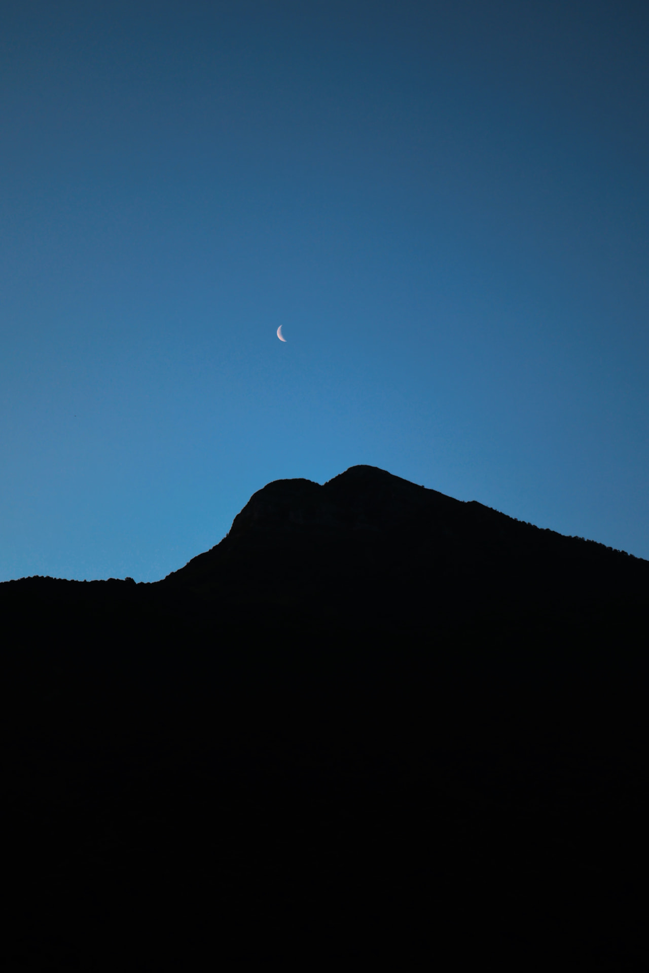 A thin crescent moon above the black silhouette of a mountain at dusk