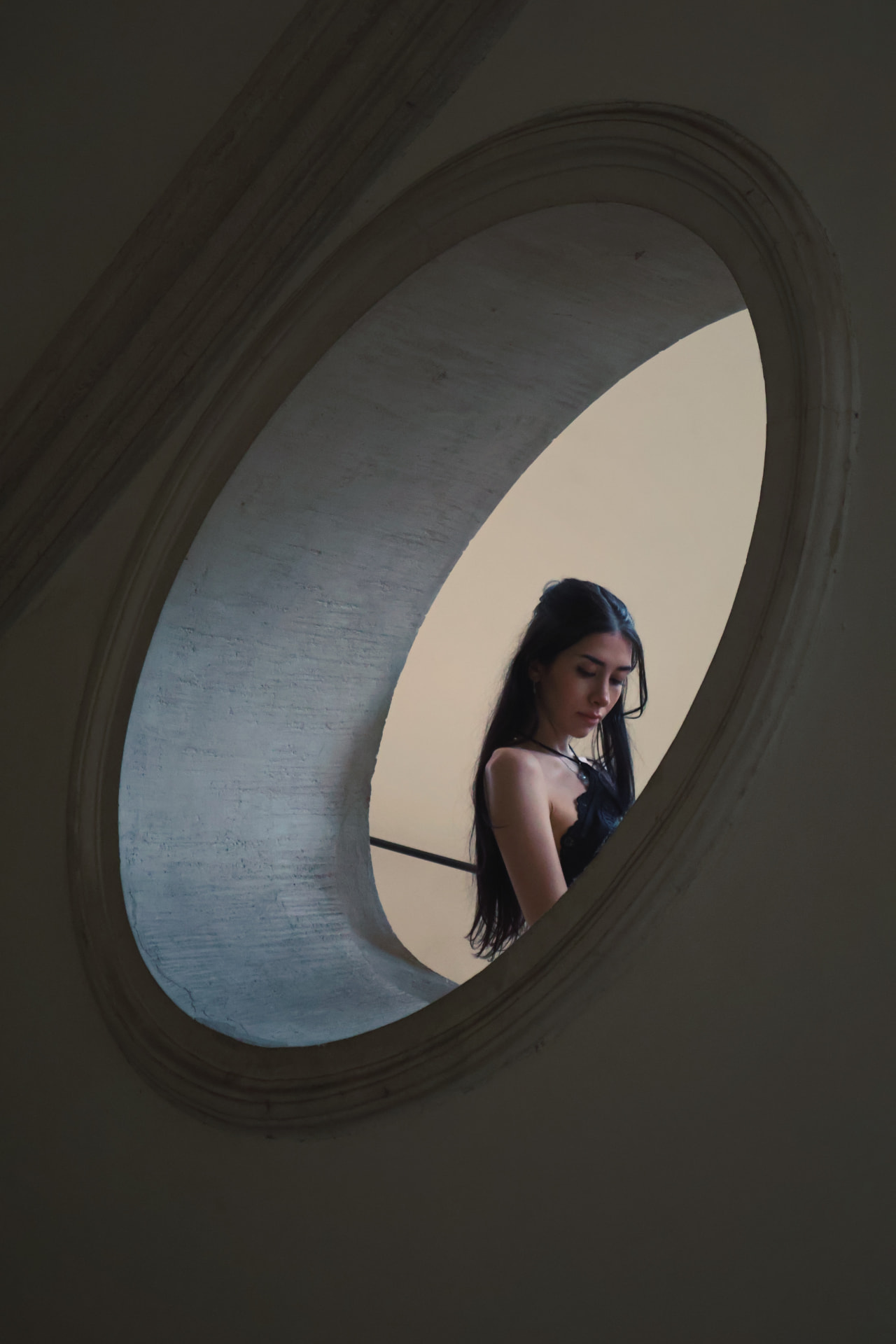 A woman seen through a tall oval window, framed by pale stone and soft daylight