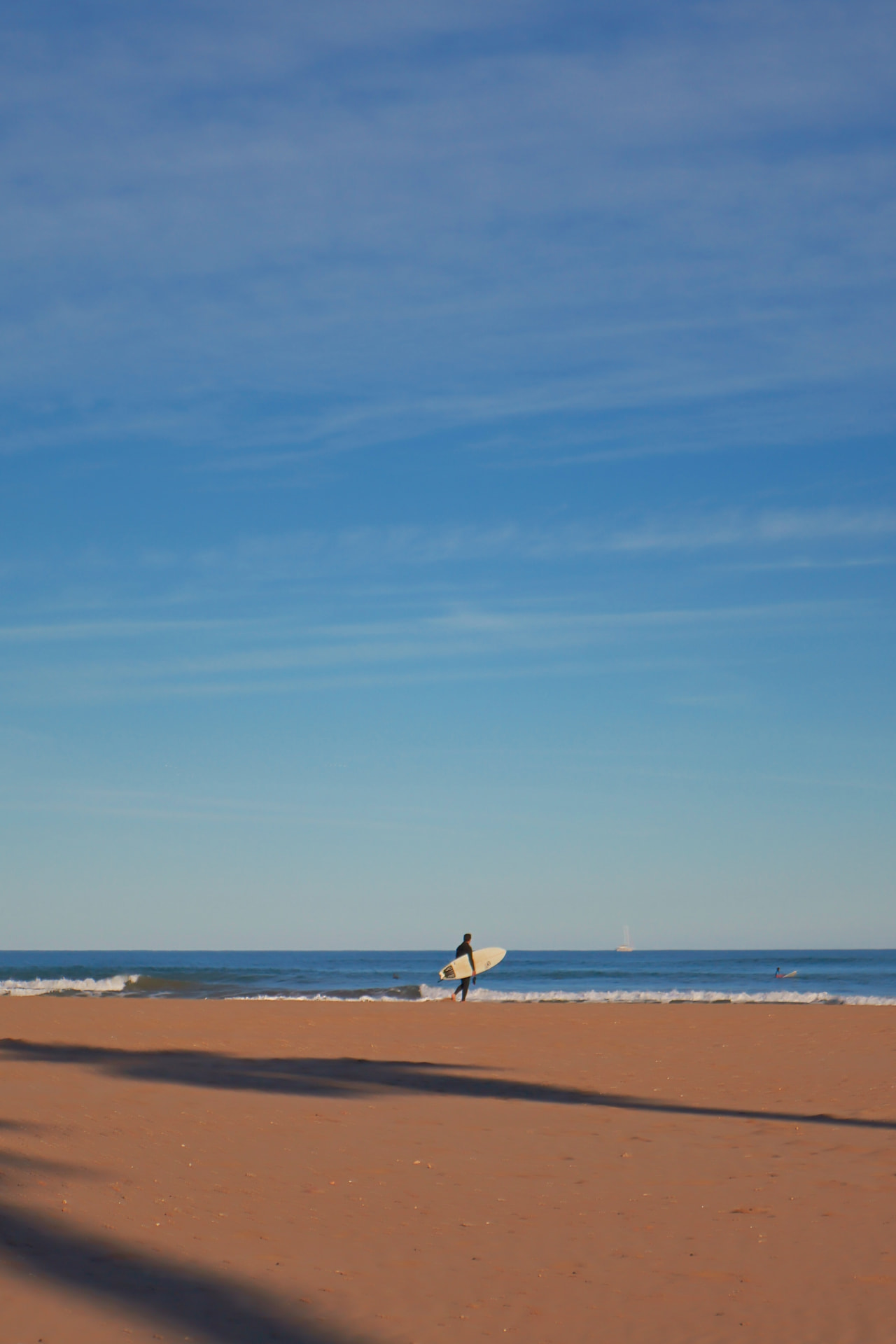 A lone surfer with a white board walking into gentle waves under a vast blue sky
