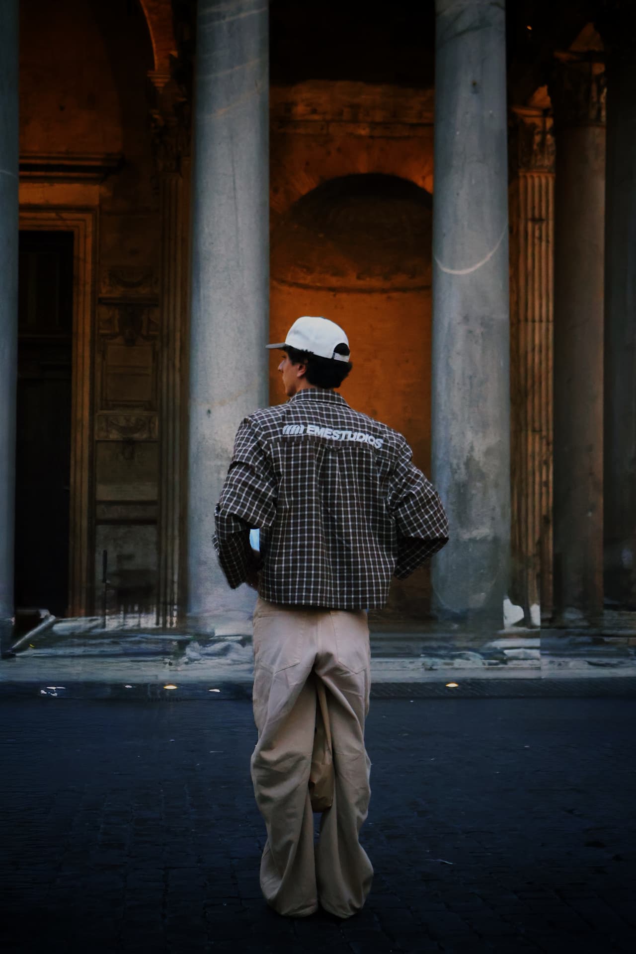 A young man in a checkered shirt standing before towering stone columns at dusk