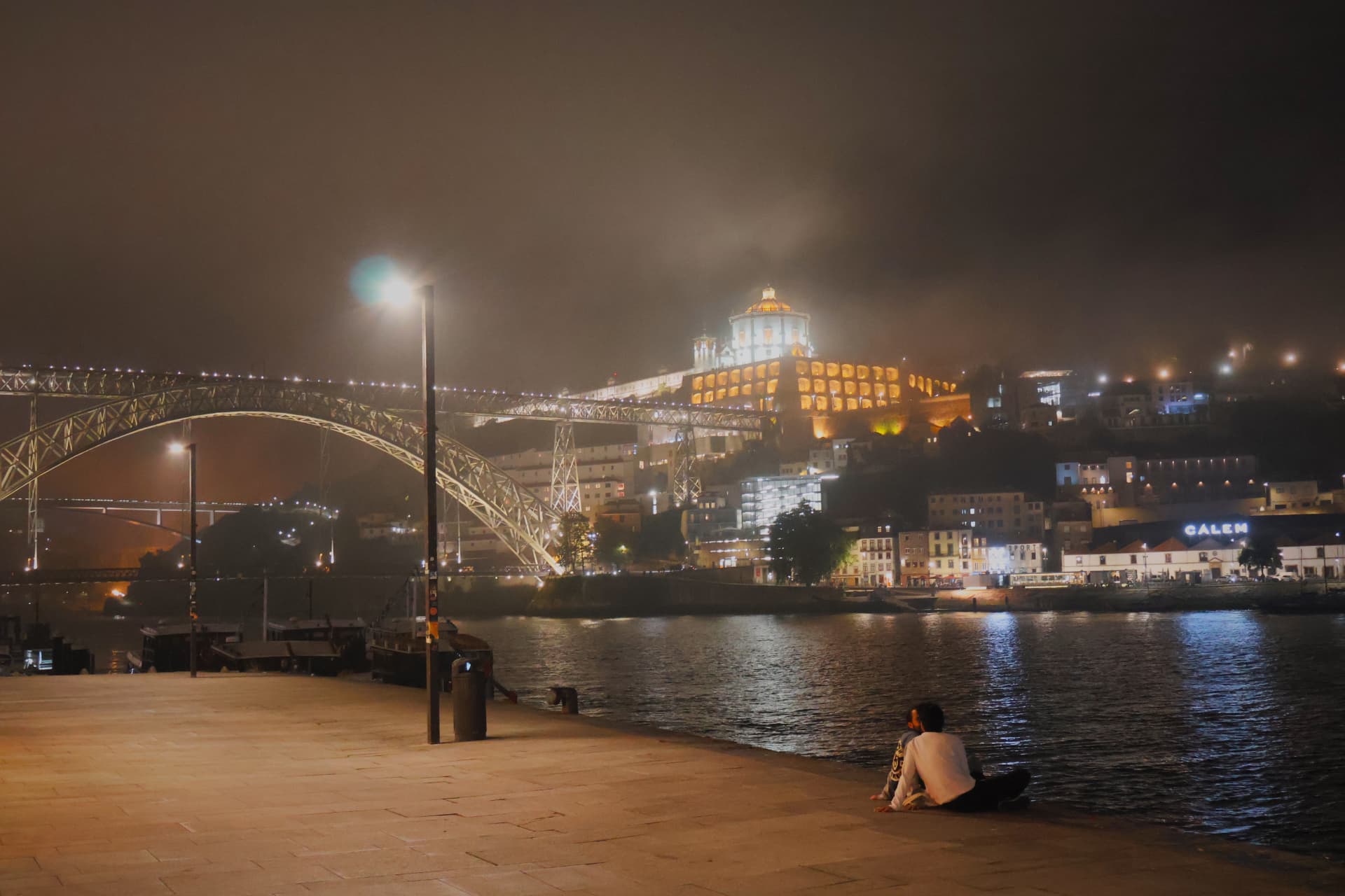 A couple sitting by the dark river at night, facing a lit bridge and hillside