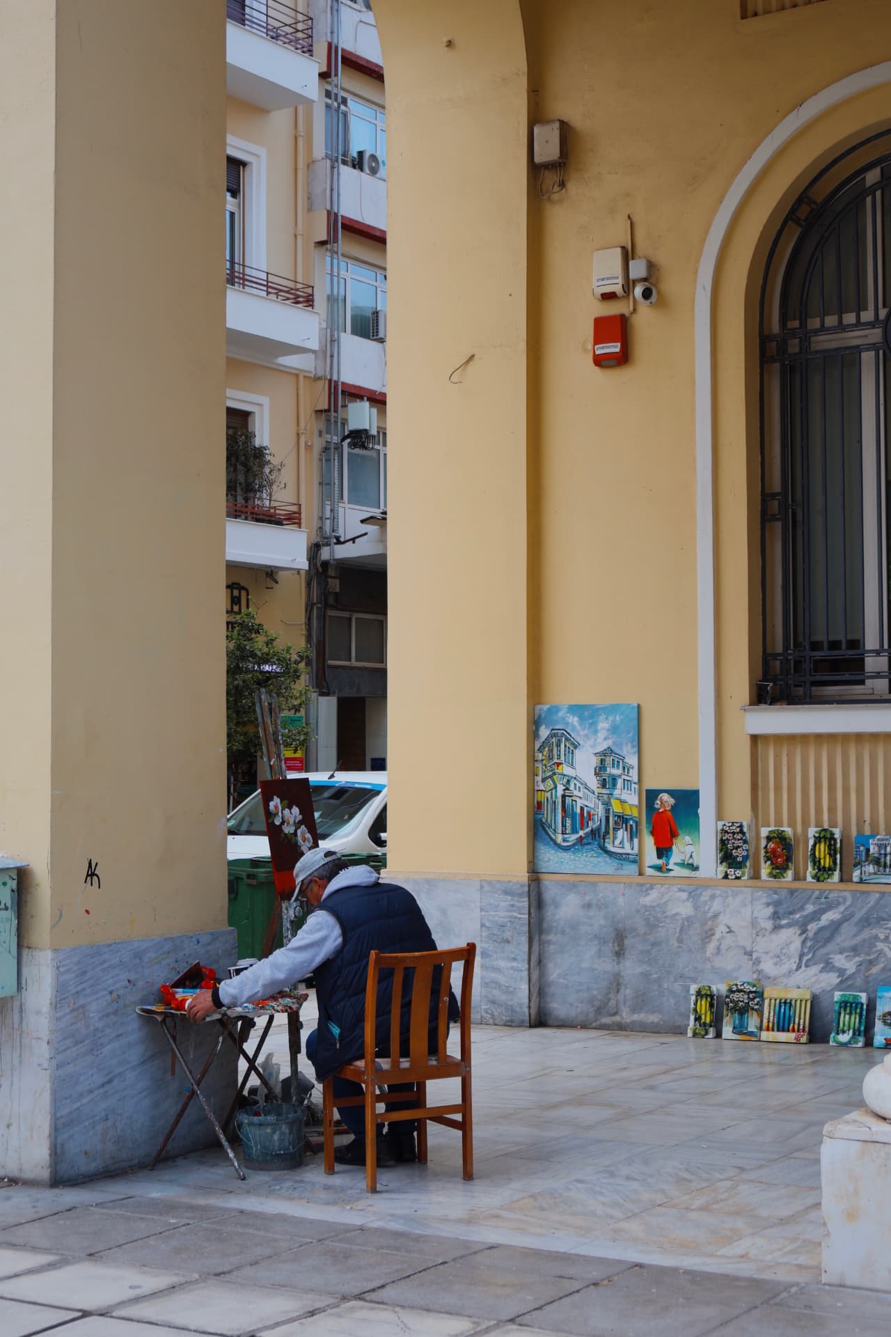 A street painter seated at his palette beneath a yellow arcade, small paintings lined along the wall
