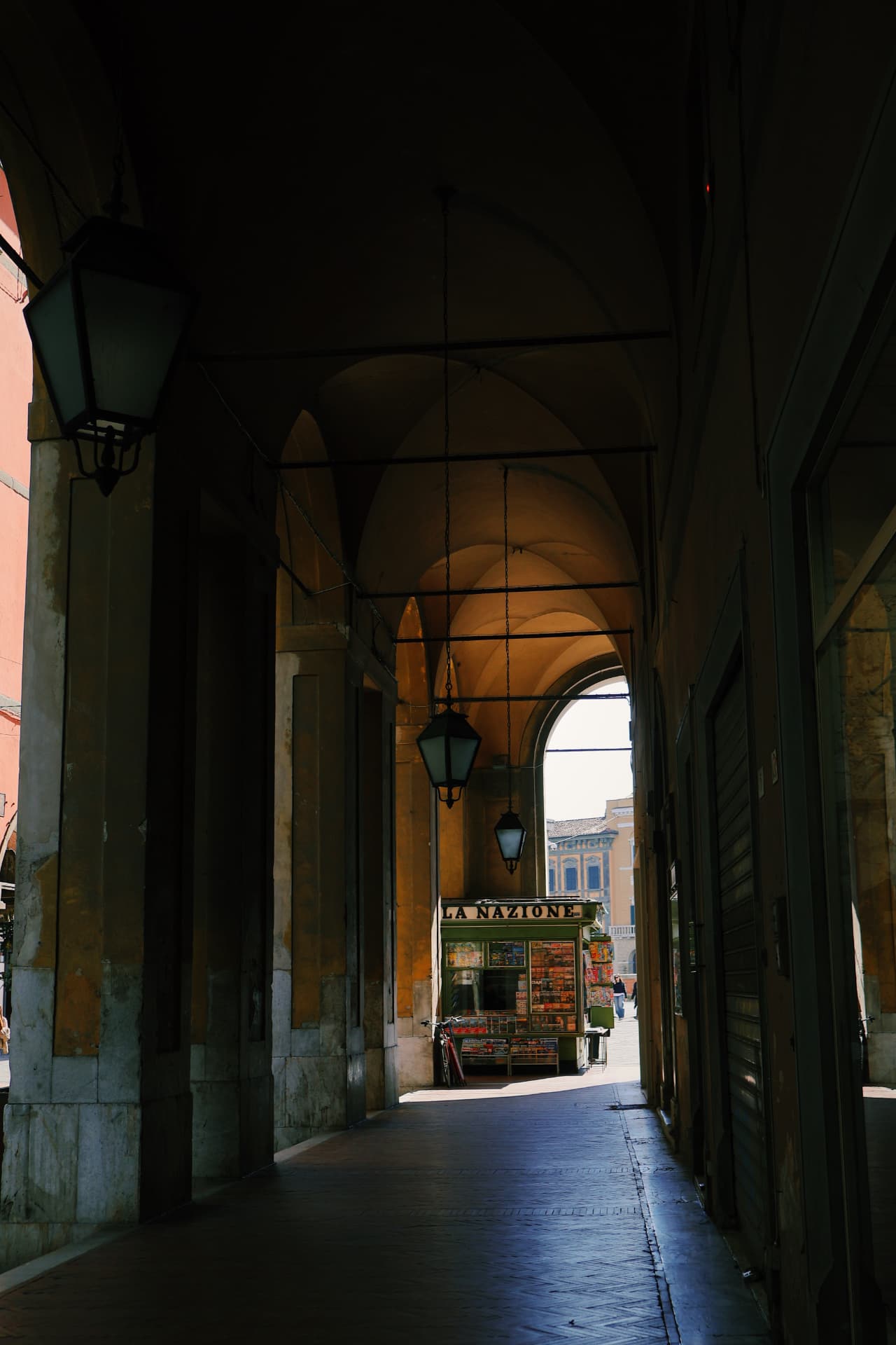 A green newsstand glowing at the bright end of a shadowed arcade with hanging lanterns
