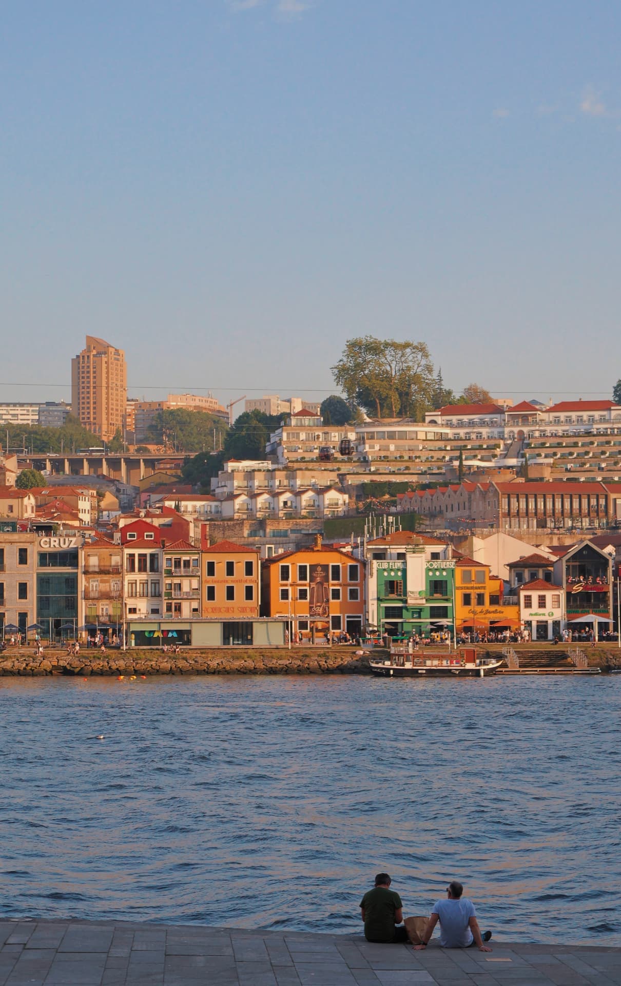Two men sitting at the river's edge facing a colourful hillside of houses